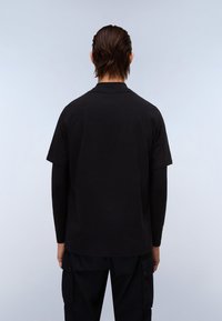 T-shirt preto de manga curta feito de algodão, com um decote redondo e um corte descontraído, usado sobre uma blusa preta de manga longa.