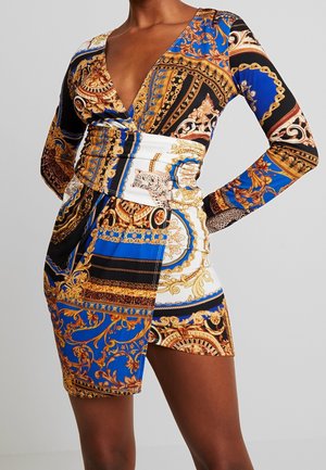 Donna che indossa un mini abito aderente a maniche lunghe con scollo a V profondo con un motivo barocco colorato in blu, oro, nero e bianco.
