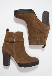 Brune ruskind ankelstøvler med en høj blokhæl, der har en ekstern lynlås, remdetalje med spænde og struktureret gummisål.