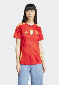 Červený fotbalový dres s žlutými akcenty a grafickými prvky, s kulatým výstřihem, krátkými rukávy a strukturovanou látkou. Logo Adidas viditelné.