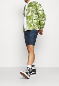 Vihreällä lehtikuviolla varustettu takki valkoisen hupparin päällä, yhdistettynä tummiin denim-shortsihousuihin ja mustiin korkeisiin tennareihin, joissa on valkoisia yksityiskohtia.