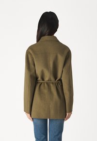 Oliivinvihreä villasekoitustakki, jossa on vyötäröllä vyö, istutettu malli ja notkollinen kaulus, nähtynä takaa. Pehmeä tekstuuri ja minimalistinen design.