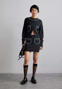 Fekete kötött kardigán és szoknya szett fehér varrott zsebekkel, hosszú ujjal és cropped stílussal. Kiegészítve áttetsző fekete térdig érő harisnyával és loaferekkel.