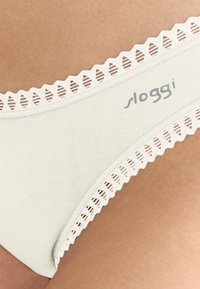 Hvidt bomuldsundertøj med bølgede blondekanter og "sloggi"-logo på hoften, båret på huden.