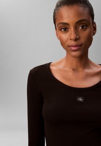Nærbillede af en kvinde med mørkt hår iført en sort Calvin Klein langærmet top mod en grå baggrund.