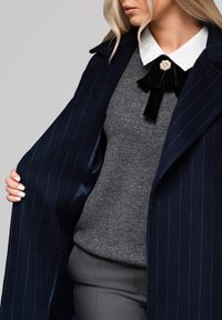 Abrigo de lana azul marino con rayas, suéter gris con un acabado texturizado y un lazo de terciopelo negro decorado con un broche de perla.