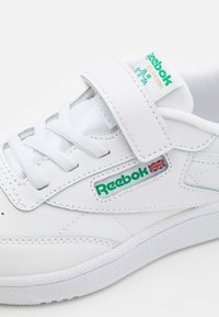 Bijele kožne tenisice s glatkom teksturom, s zelenim Reebok logotipom, bijelim vezicama i čičak trakama za sigurno prianjanje.