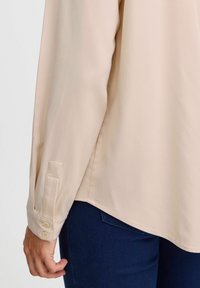 Blusa beige chiaro con maniche lunghe, polsini abbottonati e orlo arrotondato. Il tessuto ha una consistenza liscia. Indossata con jeans blu scuro.
