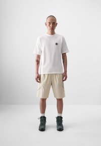T-shirt blanche à manches courtes avec logo étoile noir, associée à un short beige avec poches latérales et accents gris, complété par des bottes sombres.