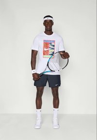 Tricou Nike alb cu un grafic colorat; pantaloni scurți negri; adidași albi; ținând o rachetă de tenis; bentiță și brățări albe.