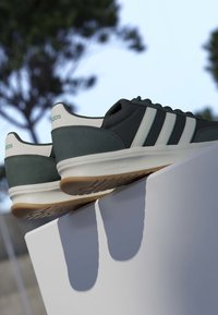 Baskets Adidas vert foncé et blanc avec semelles en gomme reposant sur une surface blanche en extérieur avec des arbres flous en arrière-plan.