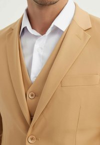 Blazer beige avec deux boutons, une petite poche poitrine et un col cranté, superposé sur une chemise blanche à col classique. Texture de tissu lisse.