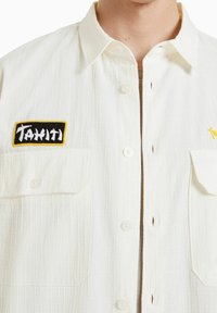 Camicia bianca con bottoni e finitura testurizzata, caratterizzata da un patch "Tahiti" nero e giallo e due tasche frontali.