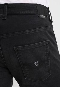 Jeans de mezclilla negros que presentan un bolsillo trasero con un parche en forma de triángulo del logo de Guess. La textura parece suave con un ligero brillo.