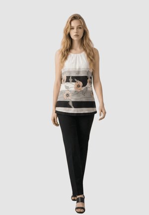 Mujer con pelo largo que lleva una camiseta blanca sin mangas con rayas negras y estampado floral, pantalones negros y sandalias negras, de pie sobre un fondo gris.