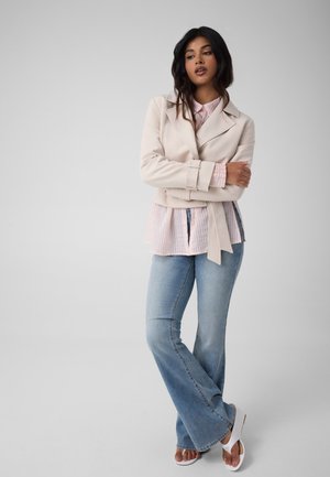 Frau mit langen dunklen Haaren, die eine beige Jacke, ein rosa gestreiftes Hemd, blaue Schlagjeans und weiße Sandalen trägt und mit verschränkten Armen steht.