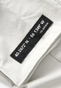 Etichetta in tessuto rettangolare nera con testo bianco che mostra le coordinate "40.2672° N - 86.1349° W" e "LTD EDITION" su tessuto beige chiaro.