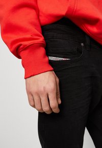 Röd sweatshirt med ribbade ärmar bredvid svarta jeans med en synlig etikett i framfickan. Handen vilar på jeansen.