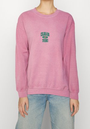 Persoon draagt een roze sweatshirt met de tekst "Colorado Springs 1995" in groen, gecombineerd met lichtblauwe spijkerbroek.