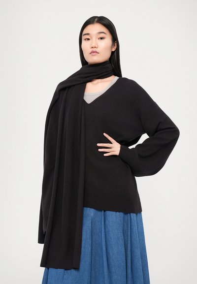 Pull en laine noir à col en V avec des manches oversized, superposé sur un haut gris et associé à une jupe plissée bleue. Écharpe drapée autour du cou.