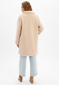 Beige, flauschiger Außenmantel mit Kragen, langen Ärmeln und lockerer Passform. Kombiniert mit hellblauen, weiten Jeans und hellbraunen Stiefeletten.