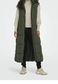 Woman wearing a long, sleeveless olive green puffer vest, beige cable knit sweater, black pants, and beige sneakers, standing against white background.