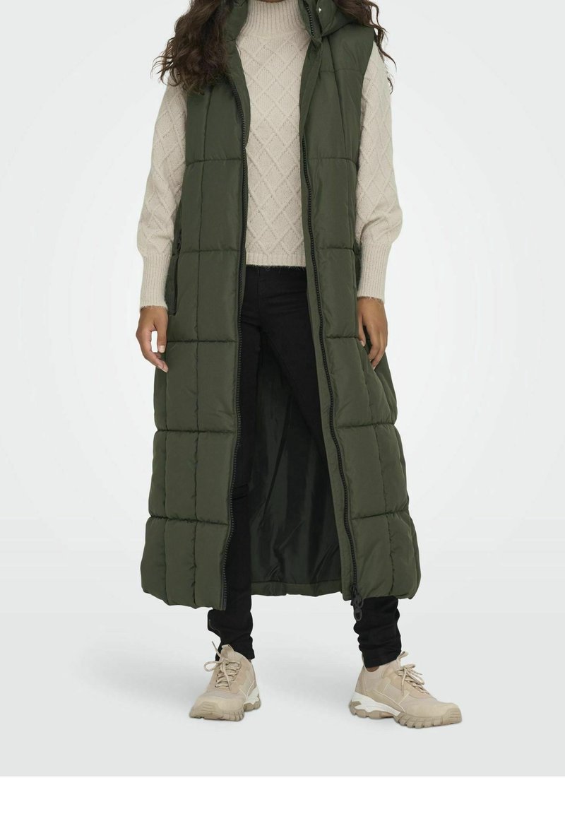 Woman wearing a long, sleeveless olive green puffer vest, beige cable knit sweater, black pants, and beige sneakers, standing against white background.