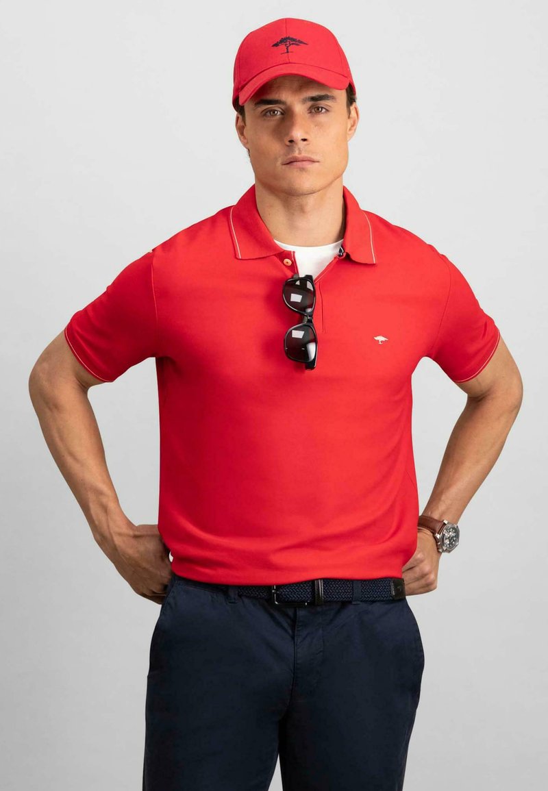 Rød polo skjorte med hvite detaljer, svarte solbriller, marineblå bukser og en matchende rød caps med logo. Glatt tekstur og klassisk design.