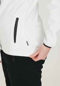 Vit softshelljacka med svarta dragkedjor och detaljer, med en ficka med dragkedja och elastiska ärmslut, samt svart byxor.