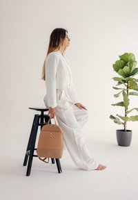 Hnědý kožený batoh zaobleného tvaru, se zipovým uzávěrem a detailem loga. Modelka v bílém oblečení sedí na černé stoličce vedle květináče.