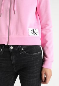 Roze zip-up hoodie van zacht materiaal met een geribbelde zoom, voorzien van een wit label met zwarte tekst. Gekleed met donkere denim jeans.