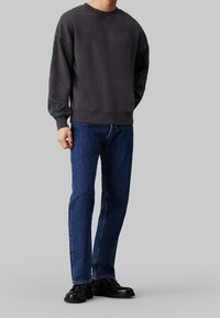 Hombre vestido con sudadera gris oscura, vaqueros azules y zapatos de cuero negros, de pie contra un fondo gris claro liso.
