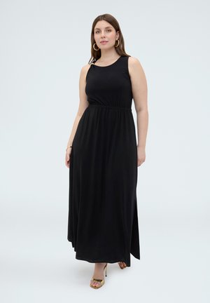 Donna che indossa un maxi abito nero senza maniche con uno spacco alto, orecchini a cerchio dorati, braccialetto d'oro e tacchi aperti dorati, in piedi contro uno sfondo bianco.