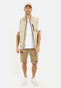 Beige mouwloze vest met zwarte rits, gecombineerd met bruin cargoshorts voorzien van zakken. Een wit shirt eronder en witte sneakers.