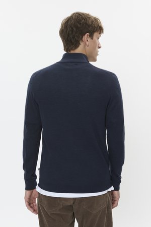 Polo de manga larga en color marino con un cuello acanalado, hecho de una tela suave. Presenta un bajo que es ligeramente más largo en la parte de atrás, combinado con pantalones de pana.