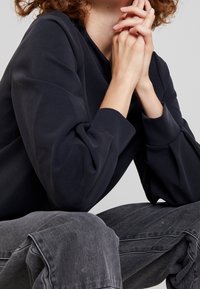 Pullover blu navale con maniche plissettate, realizzato in un tessuto morbido. Il modello indossa jeans a vita alta grigi chiari con un sottile motivo a righe.