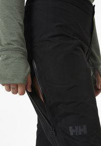 Czarne ocieplane spodnie z bocznym zamkiem, zaprezentowane w matowym wykończeniu z delikatnym logo na dolnej części nogawki. Widoczne są długie zielone rękawy.