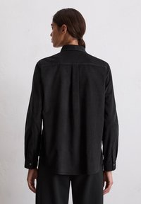 Frau mit dunkelhaarigen, nach hinten gebundenen Haaren trägt ein lockeres schwarzes Langarmshirt und schwarze Hosen und steht vor einer schlichten weißen Wand.