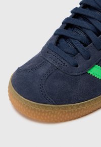 Tengerészkék velúr sportcipő zöld kiegészítővel, texturált gumitalppal és fűzős kialakítással. A közeli kép a varrás részleteit és az anyag textúráját mutatja.