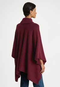 Γυναίκα που φοράει μια χαλαρή, μπορντό πλεκτή πόγκντσο με ψηλό γιακά, στεκόμενη πλευρικά μπροστά σε απλό λευκό φόντο.