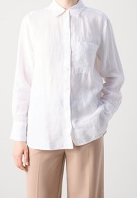 Witte linnen overhemd met knoopsluiting, gekenmerkt door een kraag, lange mouwen en een borstzak, gecombineerd met lichtbeige broeken. Zachte textuur.