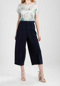Blusa ligera y transparente con un estampado abstracto en tonos verde-azul, combinada con pantalones anchos de tiro alto en color navy y tacones beige con correas.
