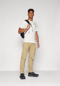 Hvit t-skjorte med oransje detaljer, beige cargo-bukser med glidelås-lommer, svart ryggsekk og svarte joggesko. Glatt tekstur av stoff.