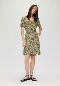 Olivegrünes Kleid mit schwarzem Blumenmuster, kurzen Ärmeln, Wickeldesign, tailliertem Bund und schwingendem Rock. Getragen mit schwarzen Riemchensandalen.