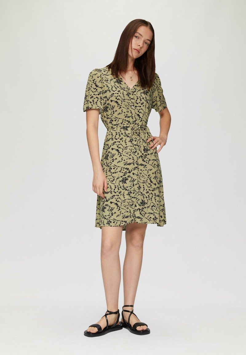 Olivegrünes Kleid mit schwarzem Blumenmuster, kurzen Ärmeln, Wickeldesign, tailliertem Bund und schwingendem Rock. Getragen mit schwarzen Riemchensandalen.