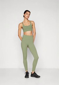 Komplet športnega oblačila: olivno zelen športni crop top in spodnji del z visokim pasom, iz gladkega, raztegljivega materiala, kombiniran s črnimi športnimi čevlji.