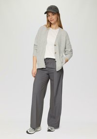 Helle graue Strickjacke mit Knöpfen, weißes T-Shirt, graue weit geschnittene Hose und graue Kappe; Model trägt metallische Sneaker.