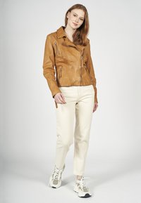 Brązowa skórzana kurtka z wyciętym kołnierzem i kieszeniami na zamek, zestawiona z kremowymi spodniami i teksturowanymi sneakersami. Gładkie tło.