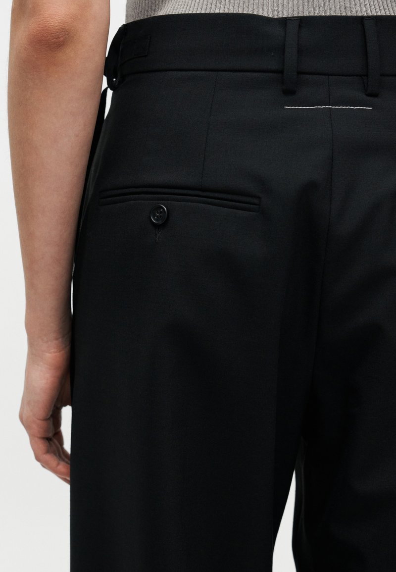Pantalon noir à texture lisse, avec une poche arrière à bouton, des passants pour ceinture et un détail de couture discret au-dessus de la poche.