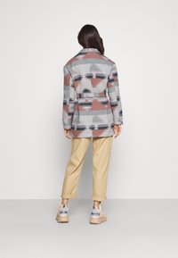 Blazer de mezcla de lana con estampado y cintura con cinturón, que presenta diseños geométricos en gris, óxido y negro. Combinado con pantalones beige y zapatillas.